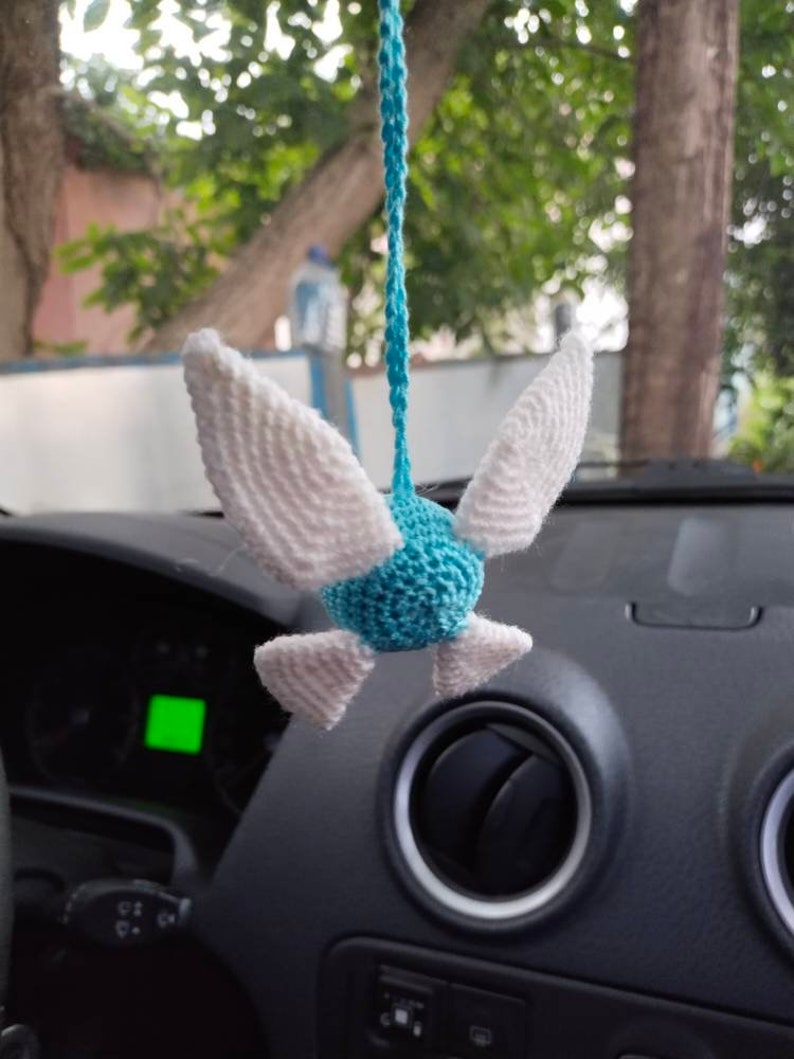 May include: A blue and white crocheted bird hanging from a rearview mirror in a car. The bird has white wings and a blue body.