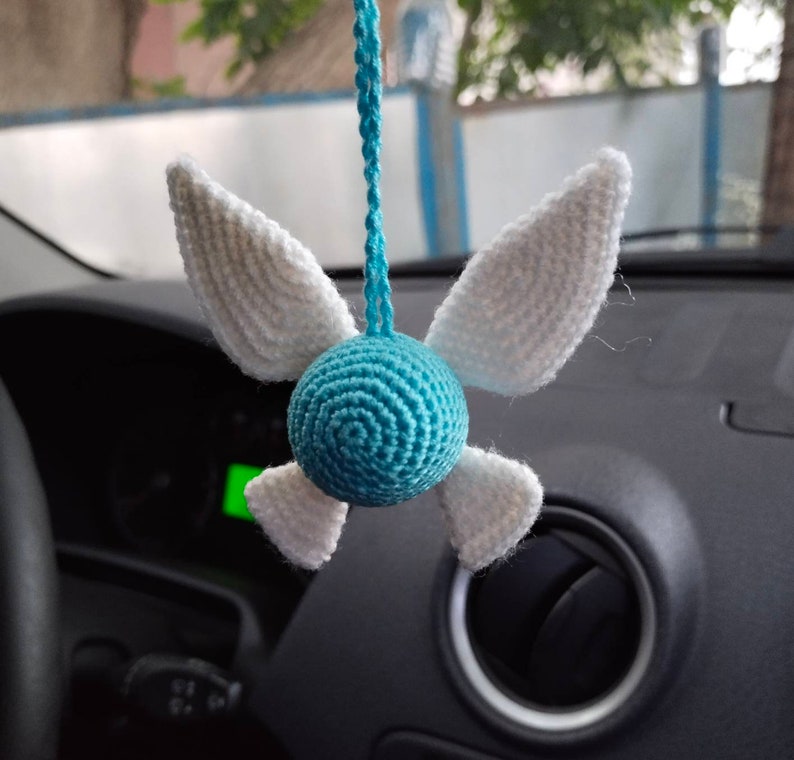 May include: A blue and white crocheted car charm shaped like a winged fairy. The wings are white and the body is blue with a circular pattern.