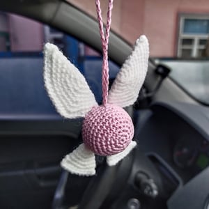 May include: A pink and white crocheted angel with wings hanging from a rearview mirror in a car.