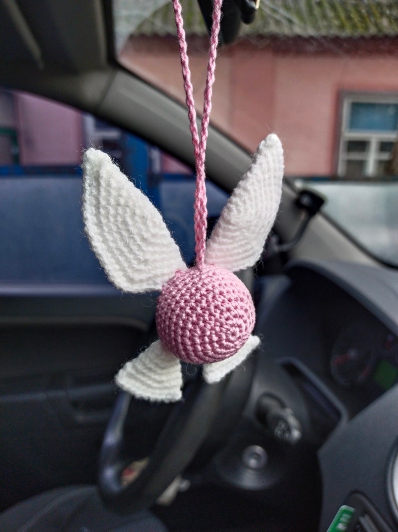 May include: A pink and white crocheted angel with wings hanging from a rearview mirror in a car.