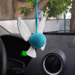 May include: A blue and white crocheted hanging ornament shaped like a bird with wings. The ornament is hanging from a rearview mirror in a car.