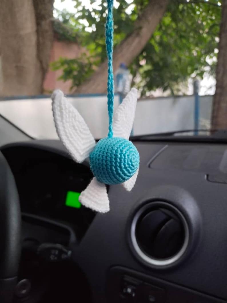 May include: A blue and white crocheted hanging ornament shaped like a bird with wings. The ornament is hanging from a rearview mirror in a car.