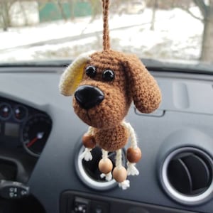 May include: A brown crocheted dog with white ears and black eyes hangs from a rearview mirror in a car. The dog has wooden beads on its legs.