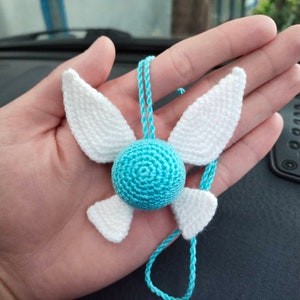 May include: A blue and white crocheted necklace with a round blue charm and white wings. The necklace is hanging from a hand.