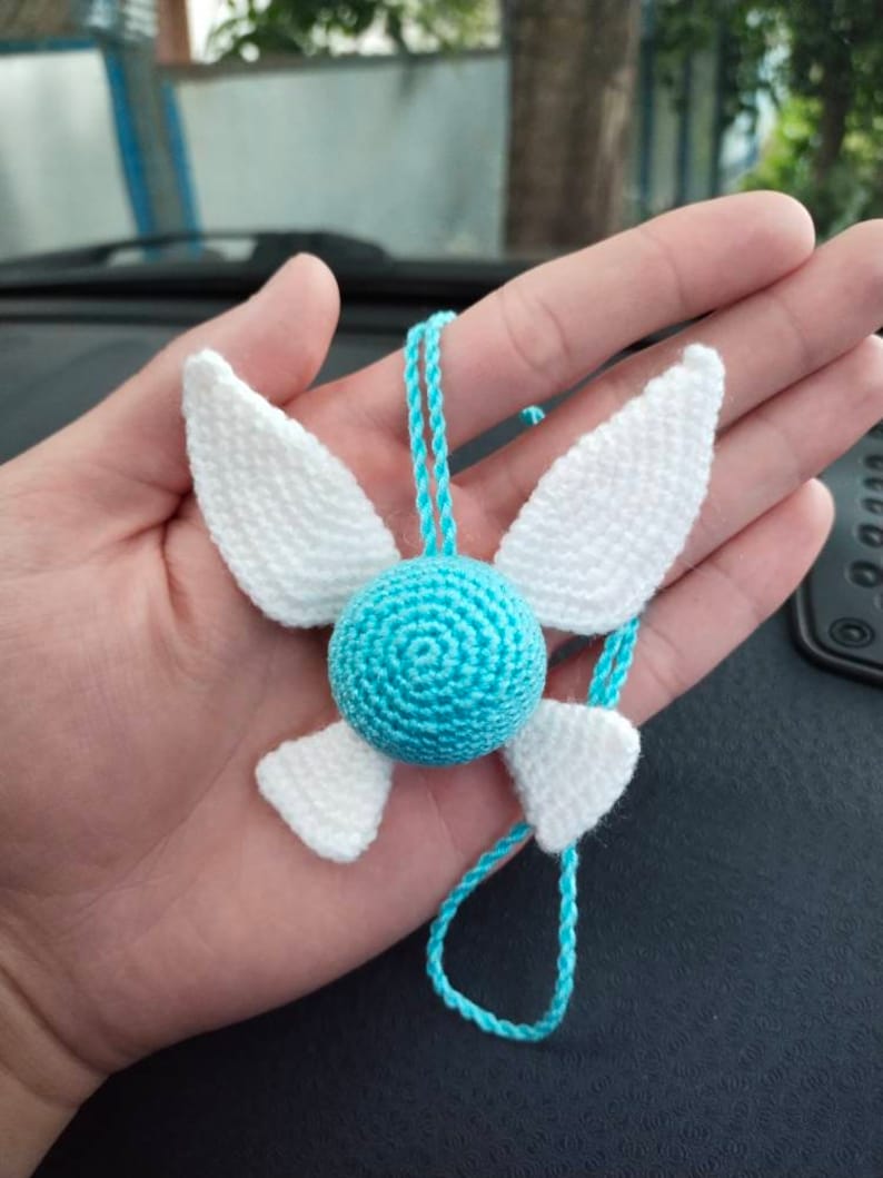 May include: A blue and white crocheted necklace with a round blue charm and white wings. The necklace is hanging from a hand.