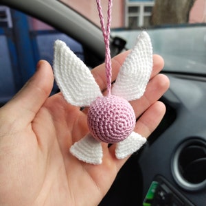 May include: A pink and white crocheted hanging ornament shaped like a fairy with wings. The ornament is hanging from a pink string.