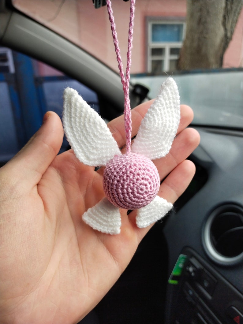 May include: A pink and white crocheted hanging ornament shaped like a fairy with wings. The ornament is hanging from a pink string.