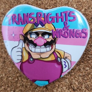 Op de afbeelding: Hartvormige button met een stripfiguur die een duim omhoog steekt. De button heeft de tekst "TRANS RIGHTS & WRONGS" in paars, tegen een achtergrond van de kleuren van de transgender pride vlag: lichtblauw, roze en wit.