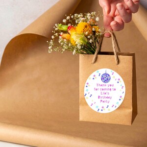 May include: A white favor bag with a round sticker featuring a colorful disco ball, stars, and musical notes. The sticker says "Thank you for coming to Lila's Birthday Party".