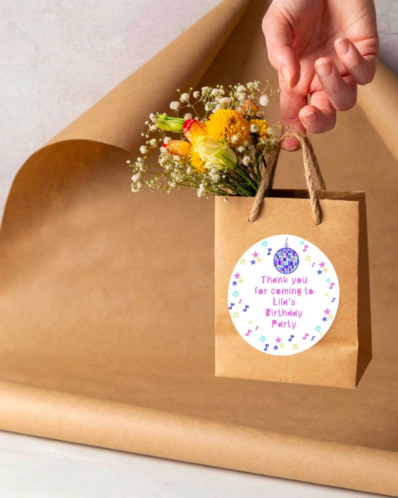 May include: A white favor bag with a round sticker featuring a colorful disco ball, stars, and musical notes. The sticker says "Thank you for coming to Lila's Birthday Party".