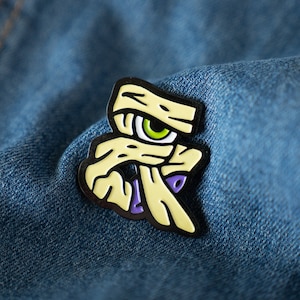 May include: A yellow and green enamel pin of a cartoon eye with bandages over it. The eye is looking to the right. The pin is on a blue denim background.
