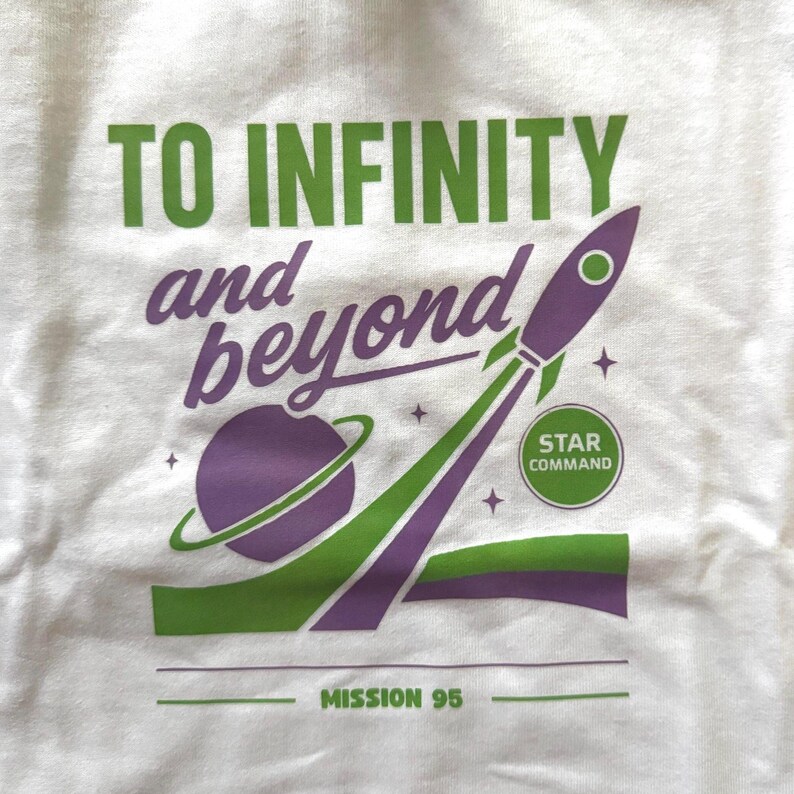 Puede incluir: Camiseta blanca con un dise&ntilde;o gr&aacute;fico verde y morado. Se ve el texto "TO INFINITY and beyond", as&iacute; como un cohete y un planeta. Tambi&eacute;n est&aacute;n las palabras "STAR COMMAND" y "MISSION 95".