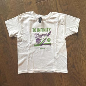 Puede incluir: Camiseta blanca con un dise&ntilde;o gr&aacute;fico que presenta un planeta p&uacute;rpura, un cohete verde y p&uacute;rpura, y el texto "TO INFINITY and beyond". La camiseta tambi&eacute;n incluye el texto "MISSION 95" y "STAR COMMAND".