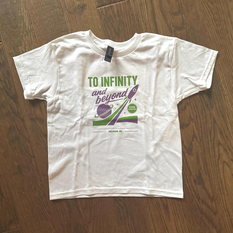 Puede incluir: Camiseta blanca con un dise&ntilde;o gr&aacute;fico que presenta un planeta p&uacute;rpura, un cohete verde y p&uacute;rpura, y el texto "TO INFINITY and beyond". La camiseta tambi&eacute;n incluye el texto "MISSION 95" y "STAR COMMAND".