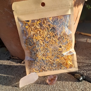May include: A brown paper pouch filled with dried yellow and purple flower petals. Three small stones are in front of the pouch: a pink stone, a brown stone, and a black stone.