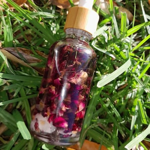 May include: A glass bottle with a bamboo lid and a dropper top filled with a dark red liquid and rose petals. The bottle is sitting on a bed of green grass.