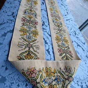 May include: Two beige fabric strips with embroidered floral patterns in shades of yellow, green, pink, and blue. The strips are folded and laid on a blue patterned surface.