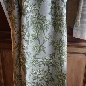 May include: A close-up of a fabric with a green and white floral pattern. The fabric is a light gray color and has a subtle sheen. The floral pattern is made up of large leaves and small flowers.