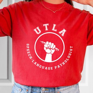 May include: Red t-shirt with a white circle design that says "UCLA Speech-Language Pathologist" with a fist holding a pencil inside the circle.