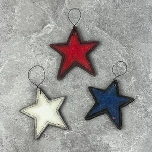May include: Three star-shaped ornaments in red, white, and blue, each with a stitched border and a wire loop for hanging. The red star is at the top, the white star is on the left, and the blue star is on the right. The ornaments are on a gray surface.