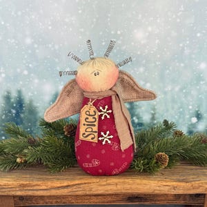 May include: A handmade angel doll with a red body adorned with candy cane and snowflake patterns. The doll has tan wings, a beige scarf, and a tag that reads "Spice". The angel's head is peach-colored with wire accents.
