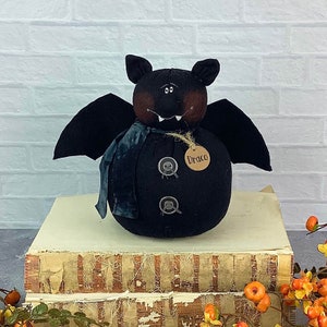 May include: A black bat-shaped stuffed toy with a brown face and large eyes. The toy has a blue scarf and two black buttons. The toy is sitting on a stack of old books and has a tag that says "Draco".