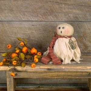 May include: A rustic autumn-themed display featuring a fabric ghost doll with a red and brown striped scarf and a tag that says "CARE". Beside it is a garland of orange pumpkins and green leaves.