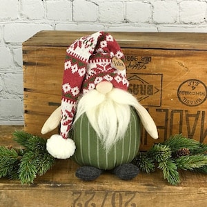 May include: A Christmas gnome doll with a long red and white patterned hat, a white beard, and a green striped body. The doll has a small wooden tag that says "Felix". The gnome is a festive holiday decoration.