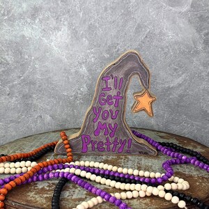 May include: Wooden witch hat decoration with the text "I'll Get You My Pretty!" in purple. A small orange star hangs from the hat. Decorative wooden beads in orange, purple, black, and white are in the foreground.