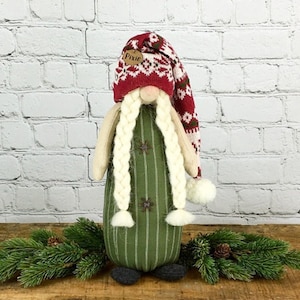 May include: A green and white striped gnome with long white braids and a red and white knit hat with the word "Pixie" on it.