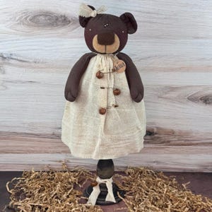 May include: A handmade brown teddy bear doll with a beige dress, wooden buttons, and a bow. The bear has a tag that says "Tessa". The doll is displayed on a black stand with a white ribbon.