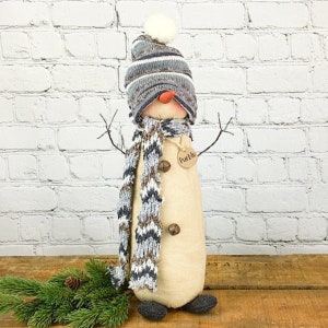 May include: A snowman figurine with a striped blue, gray, and white knit hat and matching scarf. The snowman has a carrot nose, twig arms, and a tag that says "Pueblo". The snowman is wearing a scarf with a blue and gray pattern.