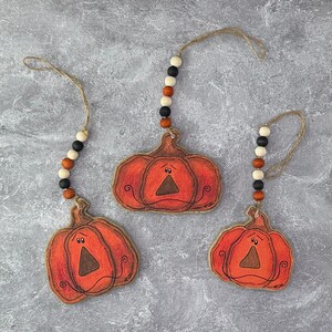 May include: Three orange wooden pumpkin ornaments with a distressed finish, each with a triangular nose and a curved mouth. They hang from twine and are adorned with wooden beads in black, white, and orange.