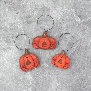 May include: Three orange pumpkin-shaped ornaments with black wire hangers. Each pumpkin has a brown stem, a triangular nose, and a smiling mouth. The ornaments are likely for Halloween or fall decor.