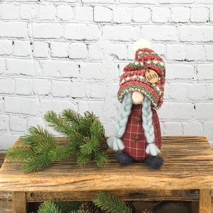 May include: A red and green plaid gnome with a white pom-pom hat and long blue braids. The gnome is wearing a wooden tag that says "Bissy".