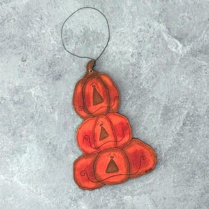 May include: A stack of five orange pumpkin ornaments with black outlines and a black string for hanging. Each pumpkin has a smiling face and a brown triangle nose. The pumpkins are arranged in a vertical stack.