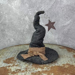 May include: A dark gray witch hat with a twisted top and a brown burlap ribbon tied around the base. A brown star is suspended from the top with a wire. The hat is sitting on a rustic, weathered surface.