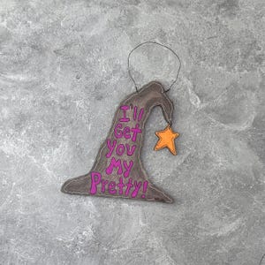 May include: A whimsical, handmade witch hat ornament with the phrase "I'll Get You My Pretty!" in pink lettering. An orange star charm hangs from the hat. The ornament is brown with black outlining and a black wire for hanging.