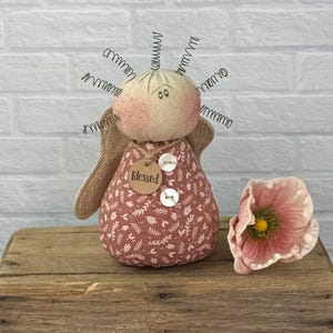 May include: A fabric angel doll with a round head, rosy cheeks, and wire halo. The doll wears a patterned dress with a tag that says "Blessed." A pink flower rests beside the doll on a wooden surface.