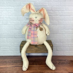 May include: A beige fabric bunny with pink ears and a plaid scarf sits on a wooden stool. The bunny has a button nose and a tag that says "Barzel".