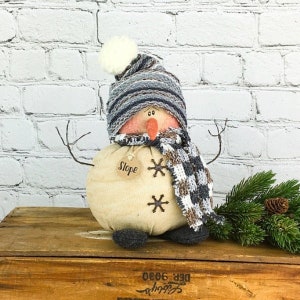 May include: A handmade snowman doll with a gray and white striped hat, a brown and white plaid scarf, and a wooden tag that says "Slope". The snowman is sitting on a wooden crate.