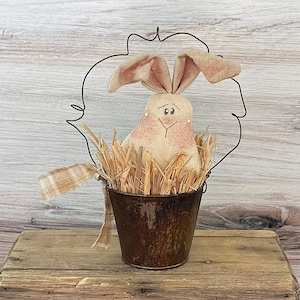 May include: A rustic Easter bunny decoration. The bunny is made of beige fabric with pink accents and sits in a rusty metal bucket filled with straw. A wire frame surrounds the bunny, and a plaid ribbon is attached to the bucket.