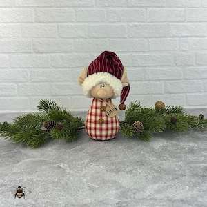 Könnte beinhalten: Eine handgefertigte Weihnachtselfenpuppe mit einem rot-weiß karierten Körper und einem passenden Hut mit weißem Besatz. Der Elf hat ein Etikett mit der Aufschrift "Bitsy". Die Puppe ist mit Tannenzweigen und Tannenzapfen dekoriert.