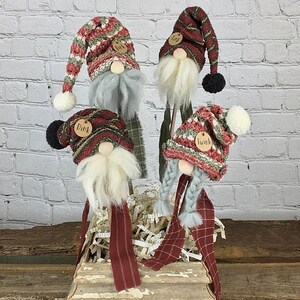 May include: Four gnome-shaped Christmas decorations with red, green, and white knit hats and scarves. The gnomes have white beards and are holding wooden sticks. The gnomes are sitting in a wooden box with shredded paper.