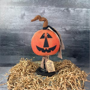 May include: A handcrafted orange pumpkin decoration with a black face, a brown stem, and a black base. The pumpkin has a tag that says "Munchkin". The decoration is placed on a bed of shredded paper, perfect for Halloween.