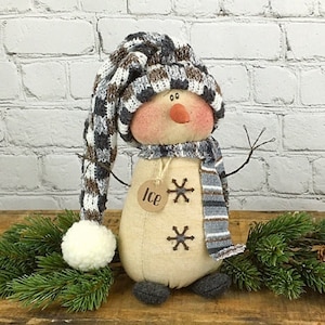 May include: A whimsical snowman doll with a long, patterned hat and scarf. The snowman has a carrot nose, twig arms, and snowflake accents. A tag with the word "Ice" hangs from its neck. The snowman is approximately 10 inches tall.