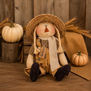 May include: A fabric scarecrow doll wearing a straw hat, a brown striped shirt, and tan overalls with a floral pattern. The doll has a beige scarf, black feet, and a small wooden tag hanging from its neck with the name Sienna printed on it.