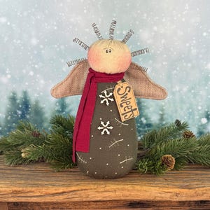 May include: A handcrafted angel doll with a round, peach-colored head and wire halo. It wears a burgundy scarf and has burlap wings. The dark green body is decorated with white snowflakes and a tag that reads "Sweets".