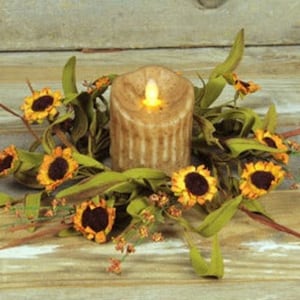 May include: A candle ring with a brown pillar candle and a flickering flame surrounded by a wreath of green leaves and yellow sunflowers.