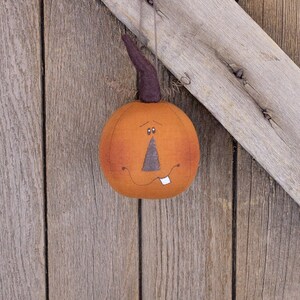 May include: A felt pumpkin ornament with a stitched-on face, hanging from a wooden surface. The pumpkin is orange with a brown stem and a toothy grin.